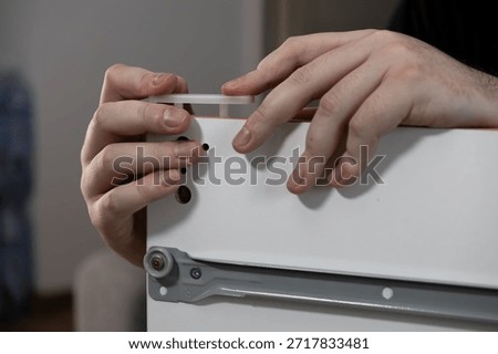The close-up view shows a person's hands installing a plastic dowel into a pre-drilled hole on a white furniture board, representing the practical process of assembling flat-pack furniture at home