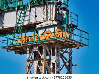 A close-up view shows the rusted structural steel framework and bright orange mechanical pulleys of a large industrial port crane against a clear blue sky. The rusted metal surfaces feature visible rivets, green railings, and metal grating platforms.