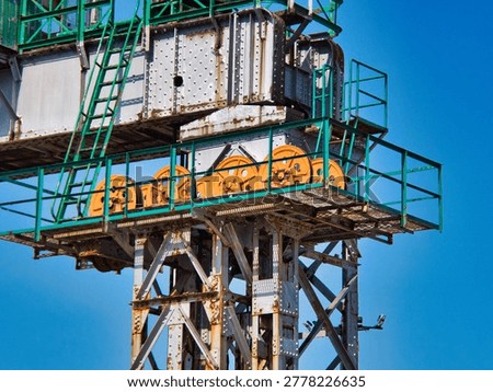 A close-up view shows the rusted structural steel framework and bright orange mechanical pulleys of a large industrial port crane against a clear blue sky. The rusted metal surfaces feature visible rivets, green railings, and metal grating platforms.