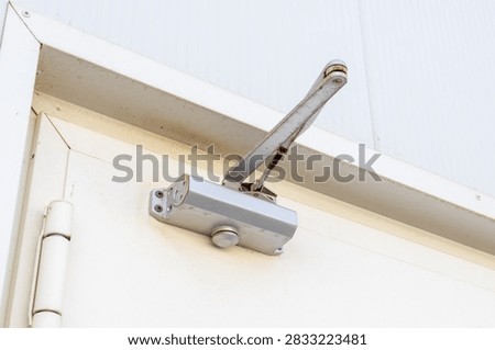 A close-up view of a silver automatic door closer attached to a white door and frame, showing its mechanical components and arm