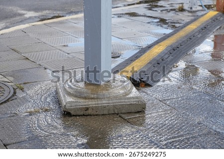 Close-up view of slick, patterned grey pavement reflecting the sky, with rainwater actively flowing around a substantial concrete pole base and a prominent safety cable ramp