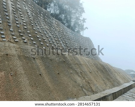 Close-up view of slope stabilization using soil nailing and shotcrete technique. Concrete square blocks arranged in a grid pattern on a steep hillside embankment to prevent landslides. Geotechnical engineering work for road safety and erosion control in Nagari Malalak, Agam. Textured concrete surface with drainage pipes visible at the base. Infrastructure repair after natural disaster showing detailed construction pattern.