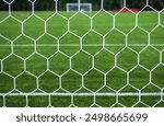 A close-up view of a soccer goal net with a lush green grass field and goalposts visible in the background.