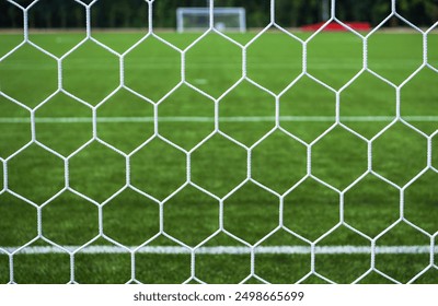 A close-up view of a soccer goal net with a lush green grass field and goalposts visible in the background. - Powered by Shutterstock