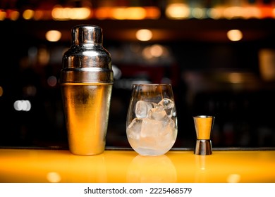 Close-up View Of Steel Shaker And Glass With Ice Cubes And Jigger On Yellow Table Surface