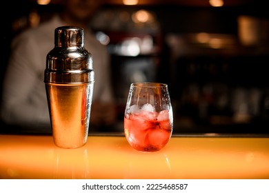 Close-up View Of Steel Shaker And Glass Of Cocktail With Ice Cubes On Yellow Table Surface