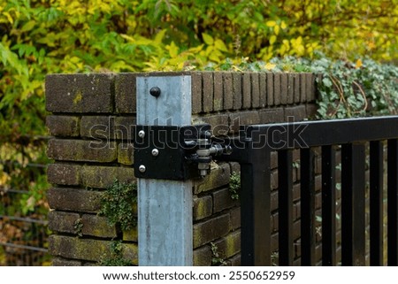 A close-up view of a sturdy black gate hinge attached to a weathered brick wall, surrounded by vibrant autumn foliage highlighting seasonal colors.