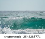 Close-up view of a teal-colored wave breaking near the shore. White foam crests the wave as it rolls in. The sunlit water sparkles. A calm ocean stretches to the horizon under a clear sky.