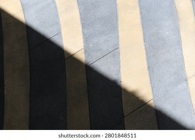 A close-up view of a textured ground surface composed of parallel strips of grey and beige paving stones intersected by a sharp diagonal shadow