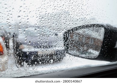 A close-up view through a car window covered in raindrops and melting snow, framing a black side-view mirror that reflects a red brick building on a wet, overcast winter day.