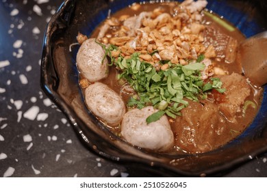 A close-up view of a traditional Thai boat noodle soup served in a black bowl, featuring tender pork meatballs, sliced pork, crispy fried garlic, and fresh herbs on top. - Powered by Shutterstock