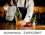 Close-up view of transparent glass with cold cocktail decorated with a sprig of mint in which bartender accurate adds carbonated liquid from siphon