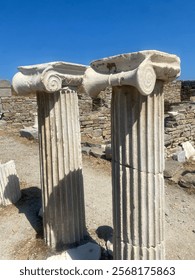 A close-up view of two ancient Ionic columns with intricate scroll-like capitals, set in a historical archaeological site under a bright blue sky. The weathered marble reflects the passage of time.