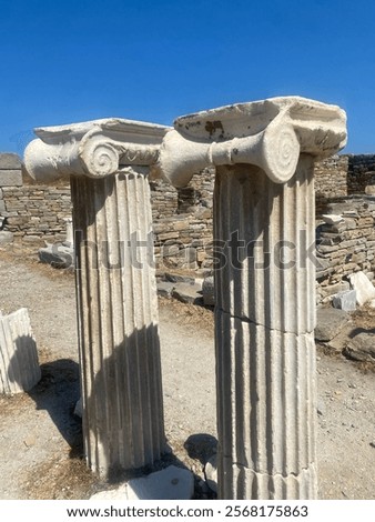 A close-up view of two ancient Ionic columns with intricate scroll-like capitals, set in a historical archaeological site under a bright blue sky. The weathered marble reflects the passage of time.