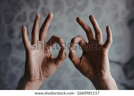 Close-up view of two hands making the okay gesture, thumbs and index fingers forming circles, against a blurred grey background.