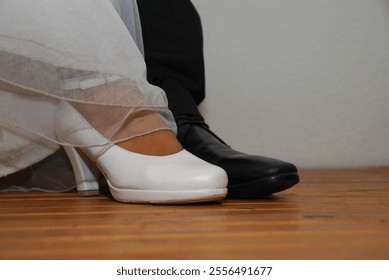 A close-up view of two shoes on a wooden floor. One shoe, the bride's, is white high-heeled , partially covered by a part of a wedding dress. The other shoe, the groom's is black. - Powered by Shutterstock