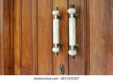 Close-up view of two vertical marble door pull handles with beige veining, mounted on classic brown wooden double doors with paneling. Includes antique brass and stainless steel fittings, plus a vintage keyhole below. Showcases traditional Indonesian tropical colonial architecture, ideal for luxury home, villa, and heritage building concepts