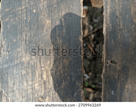A close-up view of two weathered, dark wooden planks with a gap between them. A shadow is cast across the textured surface, revealing the ground below.