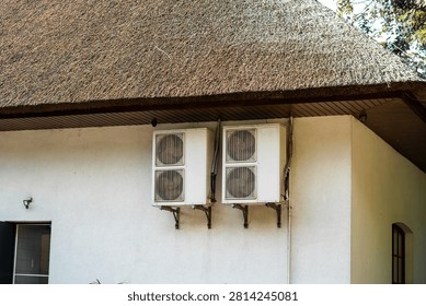 Close-up view of two white external air conditioning compressor units mounted on a light, plastered wall of a building with a dark soffit beneath a traditional, dense thatched roof, in daylight.