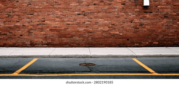 A close-up view of an urban environment featuring a textured red brick wall, a concrete sidewalk, and asphalt road with yellow parking lines.