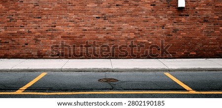 A close-up view of an urban environment featuring a textured red brick wall, a concrete sidewalk, and asphalt road with yellow parking lines.