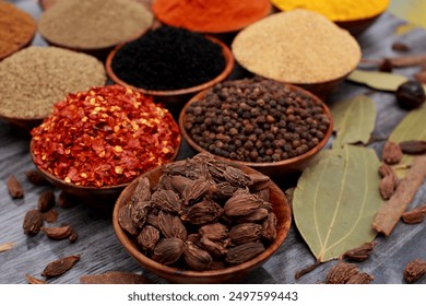 A close-up view of various spices in small bowls, including crushed red pepper flakes, black peppercorns, ground spices, and whole black cardamom pods, with bay leaves and other spices - Powered by Shutterstock