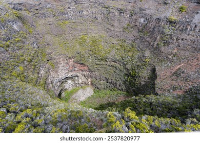 A close-up view of a volcanic crater on Piton de la Fournaise, Reunion Island. The steep, rugged walls of the crater are covered in lush vegetation, contrasting with the barren, rocky interior. - Powered by Shutterstock