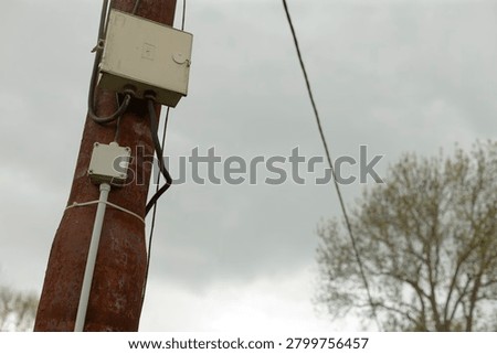 Close-up view of a weathered rusty utility pole fitted with two electrical junction boxes and insulated cables. Overhead wire stretches diagonally across a