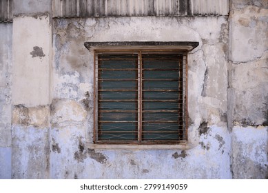 Close-up view of a weathered window with rusted metal grilles set against an aged wall with peeling paint and worn textures. The image captures the passage of time, architectural character, and urban decay commonly found in historic neighborhoods and heritage districts in Malaysia.