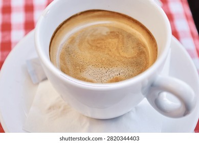 Close-up view of a white ceramic cup filled with aromatic hot espresso coffee, featuring a rich crema layer on top, served on a matching saucer with a napkin.