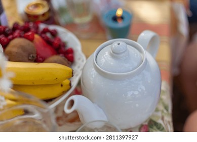 A close-up view of a white ceramic teapot next to a bowl of fresh bananas, kiwi, and cherries on a table set for an outdoor tea party or breakfas
