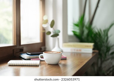 Close-up view, white coffee cup on wooden counter and smart phone, note book put on digital tablet by window, coffee break and relax after work in cafe with a reading book in the background