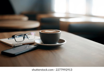 Close-up view, white cup of coffee with smartphone, notebook, pen and eye glasses on wooden table near bright window in cafe. Vintage light, blurred background - Powered by Shutterstock