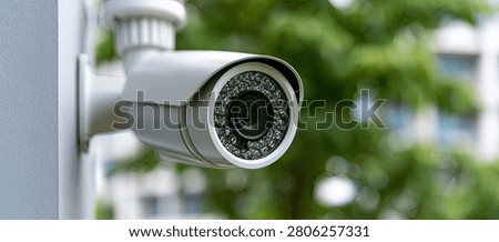 A close-up view of a white outdoor security camera attached to a grey wall, with blurred green trees and foliage visible behind it.