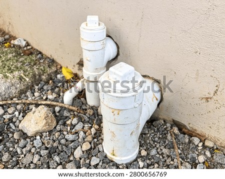 A close-up view of white PVC pipe cleanout fittings installed on the exterior wall of a residential or commercial building. The pipes, set above a gravel-covered ground, are used for plumbing maintenance and drain access in modern infrastructure.