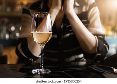 Closeup View Of White Wine Glass On Table Against Young Adult Woman Sitting On Dating At Cafe Or Restaurant At Warm Sunset Time. Alone Single Female Person Drinking Champagne At Bar