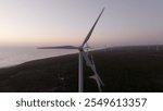 Close-up view of a wind turbine with its large blades turning, located near a coastal forest and ocean during sunset