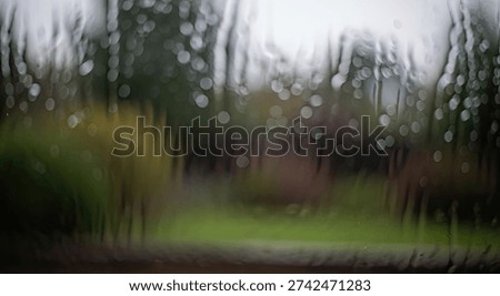 Close-up view of a window pane covered in raindrops, creating a blurred and abstract effect of the green outdoor landscape and trees beyond.
