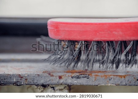 Closeup view of wire brush with red handle scrubbing rust and flaked paint from corroded metal edge, showing steel bristles, gritty residue, worn texture, and repair work in soft light