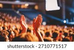 A close-up view from within a large crowd shows two hands raised high, clapping together in appreciation during a dimly lit indoor performance or presentation.