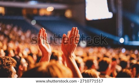 A close-up view from within a large crowd shows two hands raised high, clapping together in appreciation during a dimly lit indoor performance or presentation.