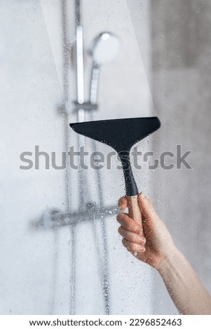 Closeup view of woman hand holding glass wiper and cleaning dirt and drops on shower partition in the bathroom. View on shower system as background. Cleaning service, washing concept