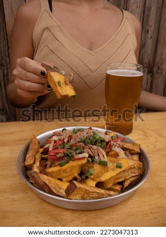 Closeup view of a woman having a beer and eating sweet potato fries with a spicy sauce made with peppers, cheddar cheese, onion and pork sausages.