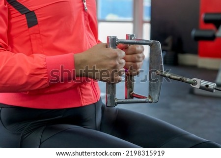 Close-up view of woman in red sportswear doing seated cable row exercise for latissimus dorsi muscles in fitness gym. Selective focus. Healthy lifestyle theme.