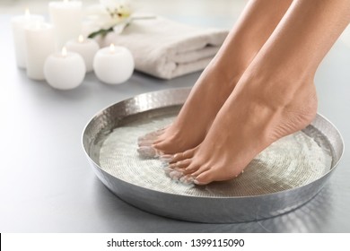Closeup view of woman soaking her feet in dish with water on grey floor. Spa treatment
