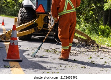 Closeup view as a worker is seen wearing high visibility clothes, using a rake to clear tree branches and leaves from road with tractor in background. - Shutterstock ID 2172652391