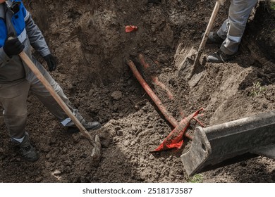 A close-up view of workers using shovels and a digger to uncover red pipes buried underground in a large dirt hole, indicating careful and collaborative excavation work.