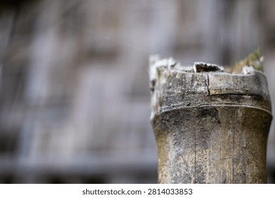 A close-up view of a worn, aged stone chimney pot with visible signs of weathering and moss growth on a blurred background with shades of gray and brown