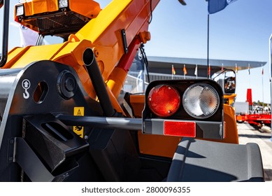 Close-up view of a yellow construction loader showing the rear light cluster, hydraulic arm, tire and part of the machine body at a busy outdoor equipment