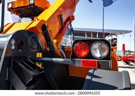 Close-up view of a yellow construction loader showing the rear light cluster, hydraulic arm, tire and part of the machine body at a busy outdoor equipment
